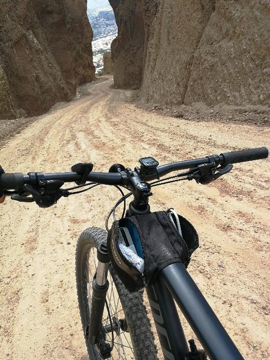 Fotografía a colores de manubrio y llanta delantera de bicicleta sobre terracería a punto de descender por camino que pasa entre dos muros de roca.