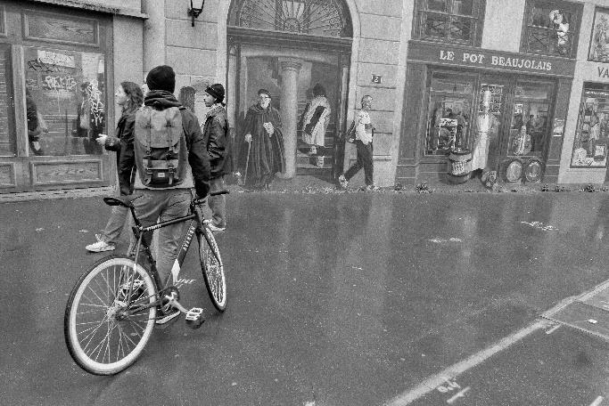 Foto en blanco y negro de escena de transeúntes que miran un mural. En primer plano, un ciclista ve la escena de espaldas al fotógrafo.