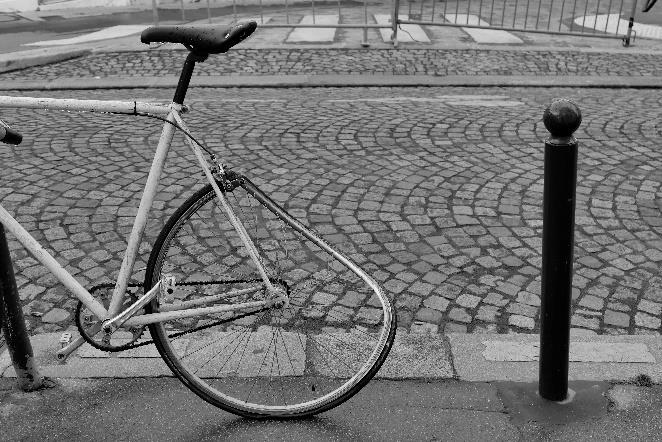 Foto en blanco y negro de llanta trasera de bicicleta deformada, de fondo se ve un suelo adoquinado.