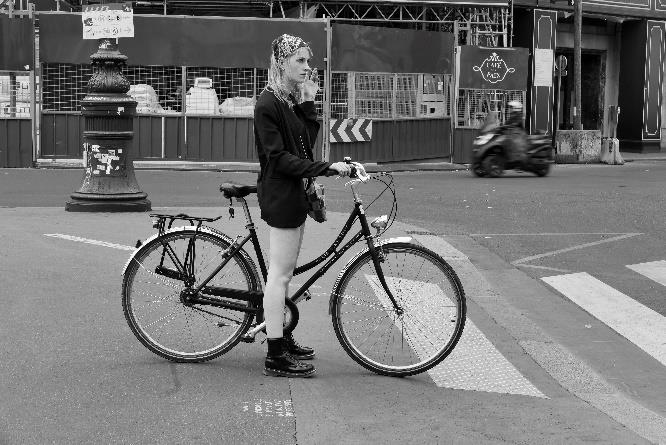 Foto en blanco y negro de mujer que viste short negro, botas industriales y paliacate en la cabeza esperando de pie sobre su bicicleta sobre la cebra peatonal para cruzar la calle.